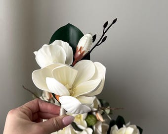May include: A close-up of a white magnolia flower with a dark green leaf and brown stems. The flower has multiple petals and is in full bloom. Other white flowers are visible in the background.