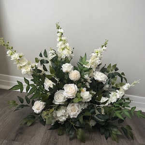 May include: A floral arrangement featuring white roses, snapdragons, and other white flowers with green foliage. The arrangement is placed on a wooden surface and against a light gray wall. The flowers are clustered together, creating a full and lush appearance.