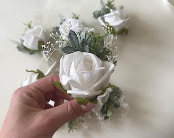 Pode incluir: Boutonniere de rosa branca com folhagem e gipsofila. A rosa é feita de tecido e a folhagem é artificial.