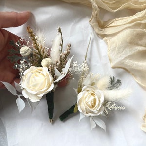 May include: Two small white flower arrangements with dried grasses and white feathers. The flowers are white roses with green stems.