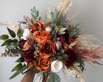 Puede incluir: Un vibrante ramo de flores con rosas naranjas, burdeos y blancas, acentuado con follaje verde y hierba de la pampa. El arreglo se sostiene sobre un fondo neutro, mostrando los ricos colores y texturas del diseño floral.