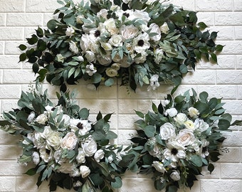 May include: Three white and green floral arrangements with white roses, anemones, and greenery. The arrangements are on a white brick wall.