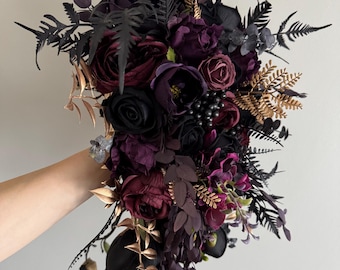 May include: A cascading bouquet featuring a mix of dark burgundy, black, and purple artificial flowers. The arrangement includes roses, peonies, and other floral elements, accented with gold and black foliage. The bouquet is held against a neutral background.
