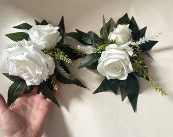 Könnte beinhalten: Zwei Ansteckblumen mit weißen Rosen, grünen Blättern und grünen Zweigen.