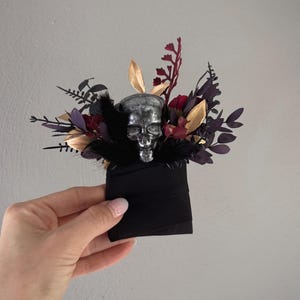 May include: A gothic-themed floral arrangement featuring a silver skull centerpiece. The arrangement includes dark red, purple, and gold-colored leaves and flowers, with black feathers. The base is a black fabric. A unique, handcrafted accessory.