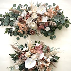 May include: Two floral arrangements featuring white calla lilies, blush-toned orchids, and dried pampas grass. The arrangements are accented with green eucalyptus leaves and other greenery, creating a natural, elegant look. Perfect for weddings or home decor.