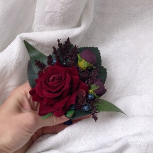 May include: A burgundy velvet rose with green leaves and dark purple berries, a small bouquet for a wedding or special occasion.