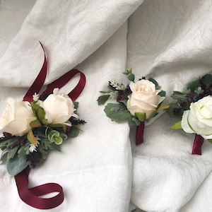 Puede incluir: Tres boutonnières de rosas blancas con cinta color burdeos y vegetación.