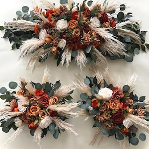 May include: A set of three floral arrangements featuring a mix of dried pampas grass, eucalyptus, and faux roses in shades of orange, peach, and white. The arrangements are perfect for a wedding or special event.