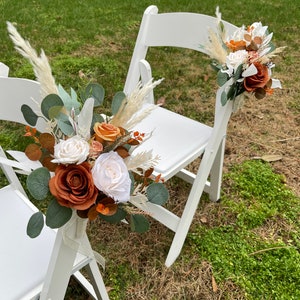 Op de afbeelding: Twee witte klapstoelen versierd met herfst-thema bloemstukken. De arrangementen bevatten gedroogd pampasgras, eucalyptusbladeren, witte en oranje rozen en andere gedroogde bloemen.
