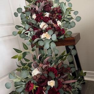 Könnte beinhalten: Zwei Blumenarrangements mit burgunderfarbenen Rosen, weißen Rosen und dunkelroten Dahlien, akzentuiert mit grünen Eukalyptusblättern. Die Bouquets sind auf einem dunklen Holzschemel angeordnet und bilden eine ansprechende Präsentation.
