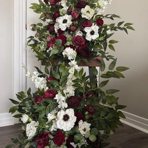 May include: A two-tiered floral arrangement featuring burgundy roses, white anemones, and greenery. The flowers are arranged on a dark wooden stand, creating a visually appealing display. The arrangement is suitable for weddings, events, or home decor.