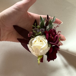 May include: A small bouquet of artificial flowers with a white rose, burgundy roses, and green leaves.