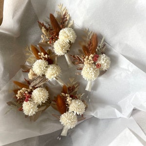 May include: Four small bouquets of dried flowers with white blossoms and brown dried grasses. The bouquets are arranged on a white surface.