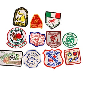 May include: A collection of 12 colourful embroidered patches featuring football themes. The patches feature various logos, including team names, mascots, and footballs. Some patches have text, such as "T-B-A-Y-S", "Pine & Rose", "International Soccer Classic", "Agincourt Soccer Club", "Streetsville Meadowvale", "Hamilton Sparta", "Erin Mills Soccer Club", "North East London Soccer Club", "Maple Leaf Soccer Club", "Oakville Soccer Club", and "International Tournament".