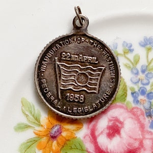 May include: Silver charm commemorating the inauguration of the West Indies Federal Legislature on 22nd April 1958. The charm features a stylised flag with the text "22nd APRIL 1958".