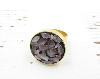 May include: A gold-toned ring with an oval face filled with small, irregularly shaped, light purple stones. The stones are set against a dark background, creating a contrast. The ring is on a white surface.
