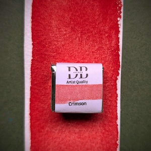 Könnte beinhalten: Ein kleines quadratisches Aquarellfarben-Pfännchen mit der Aufschrift DD Paints Artist Quality Crimson, zentriert auf einem vertikalen Papierstreifen mit einem leuchtend karmesinroten Farbmuster, das über und unter dem Pfännchen auf dem Papier verläuft.
