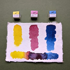 May include: Watercolor paint swatches in yellow, violet, and blue on textured paper. Three small boxes of paint are above the swatches. The boxes are labeled with the paint color names. The swatches show color gradients.