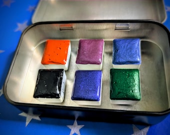 May include: A silver metal tin containing six vibrant watercolor paint blocks. The colors include orange, purple, blue, black, dark blue, and green. Each block is rectangular and sits in a white plastic base.