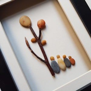 May include: A framed pebble art piece depicting a tree with a round, light brown top and a brown branch with small, round, orange and yellow pebbles.  A line of six gray, orange, and yellow pebbles are arranged at the bottom of the frame, resembling a family.