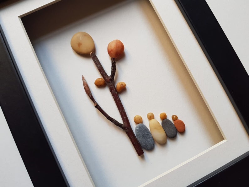 May include: A framed pebble art piece depicting a tree with a round, light brown top and a brown branch with small, round, orange and yellow pebbles.  A line of six gray, orange, and yellow pebbles are arranged at the bottom of the frame, resembling a family.