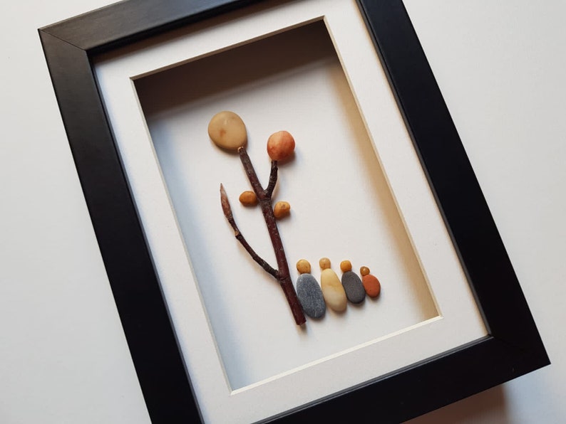 May include: A framed pebble art piece depicting a tree with a brown branch and orange and yellow pebbles representing leaves and fruit.  Five smaller pebbles in shades of gray, yellow, and orange are arranged at the base of the tree, resembling a family.