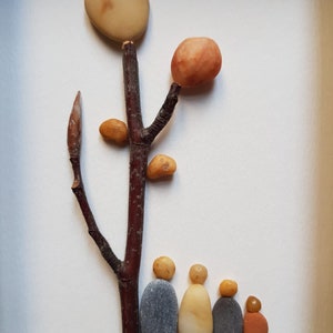 May include: A stone art piece depicting a tree with a family of five standing beneath it. The tree is made of a brown twig with a round, light brown stone at the top and smaller, orange and brown stones for leaves. The family is made of five stones, two gray, two light brown, and one orange.