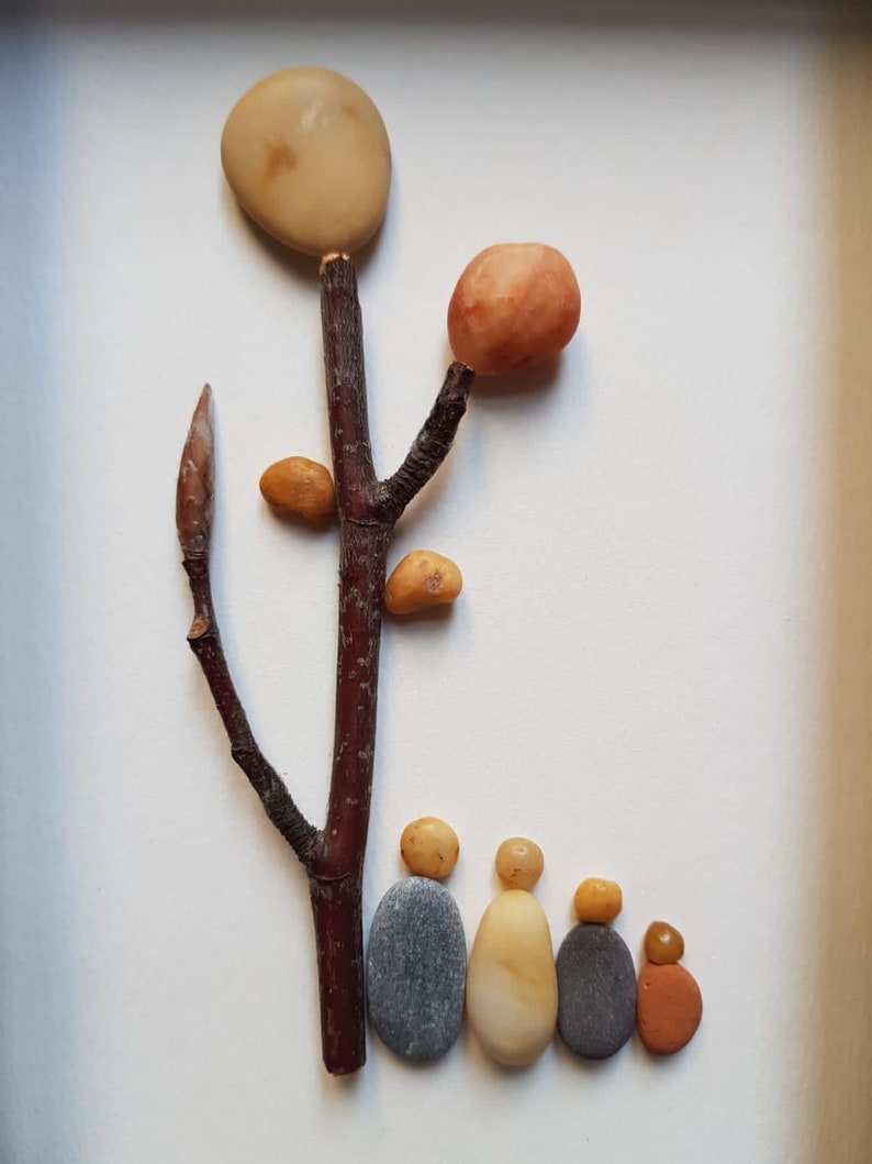 May include: A stone art piece depicting a tree with a family of five standing beneath it. The tree is made of a brown twig with a round, light brown stone at the top and smaller, orange and brown stones for leaves. The family is made of five stones, two gray, two light brown, and one orange.