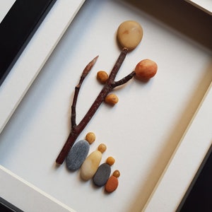 May include: A framed artwork featuring a tree made of small stones and twigs. The tree has a round, light brown stone at the top, and several smaller stones in orange and yellow shades. Five gray and yellow stones are arranged at the base of the tree, resembling a family.