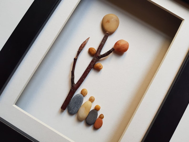 May include: A framed artwork featuring a tree made of small stones and twigs. The tree has a round, light brown stone at the top, and several smaller stones in orange and yellow shades. Five gray and yellow stones are arranged at the base of the tree, resembling a family.