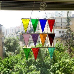 Puede incluir: Un atrapasoles de vidrieras de colores con paneles triangulares en varios colores, incluyendo amarillo, verde, azul y rojo. Los paneles están dispuestos en un diseño escalonado y suspendidos por cadenas, creando una vibrante decoración de ventana.