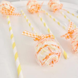 May include: Several cake pops on a white surface. Each cake pop is wrapped in clear cellophane and tied with a gingham ribbon. The cake pops have yellow and orange sprinkles and are attached to yellow and white striped paper straws.
