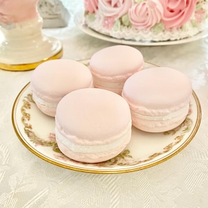 May include: Four pastel pink macarons with white filling are arranged on a decorative plate with a gold rim and floral design. A cake with pink frosting and roses is in the background. The scene is set on a white tablecloth.