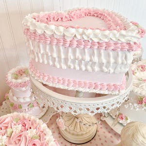 May include: A heart-shaped cake with pink and white frosting, displayed on a white, ornate cake stand. The cake features ruffled frosting and decorative pink accents. Other cakes with floral decorations are also visible.