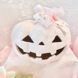 May include: A white ceramic jack-o'-lantern pumpkin with a carved face featuring triangular eyes, a triangular nose, and a wide jagged mouth. The pumpkin is decorated with a sheer pink ribbon and a small floral fabric bow on top.
