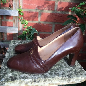 May include: A pair of brown leather heeled shoes with a gold buckle detail. The shoes have a pointed toe and a closed back.