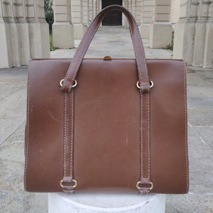 Puede incluir: Un maletín de cuero marrón con asa superior y dos correas. El maletín tiene un diseño cosido y herrajes dorados.