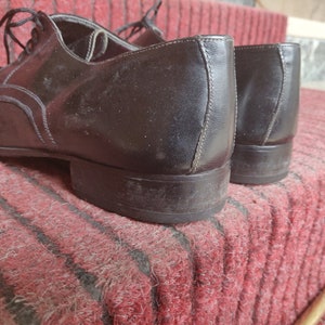 May include: A pair of black leather dress shoes with laces, resting on a red and black striped rug. The shoes have a classic design with decorative stitching and a low heel.