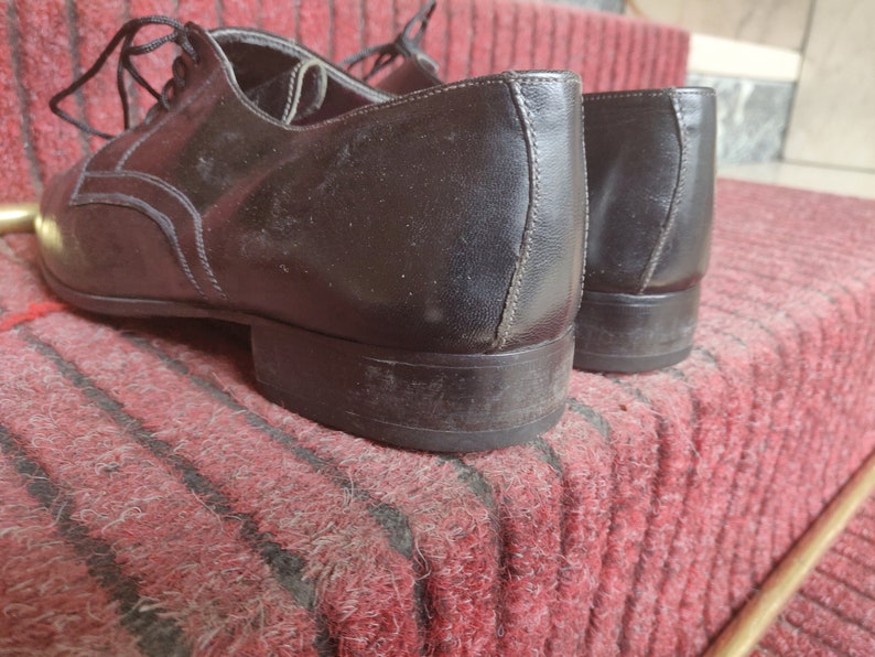 May include: A pair of black leather dress shoes with laces, resting on a red and black striped rug. The shoes have a classic design with decorative stitching and a low heel.
