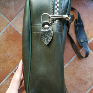 May include: A dark green leather satchel bag with a zipper closure and a silver metal clasp. The bag has a green trim and a long strap.