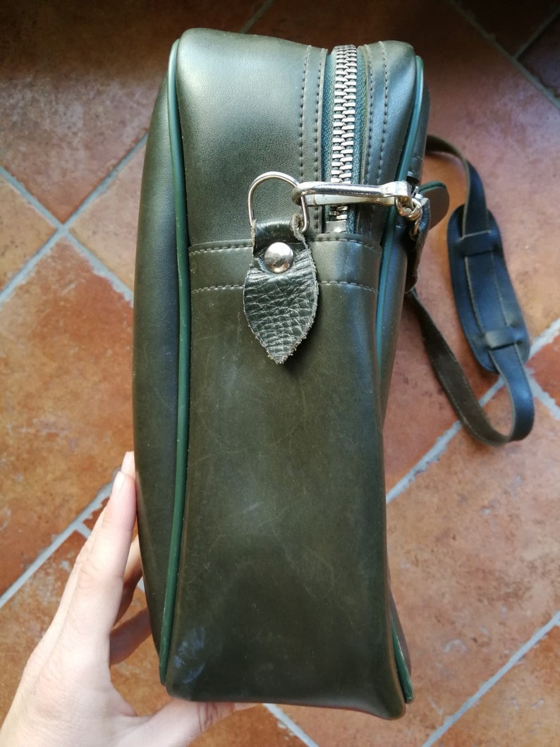 May include: A dark green leather satchel bag with a zipper closure and a silver metal clasp. The bag has a green trim and a long strap.