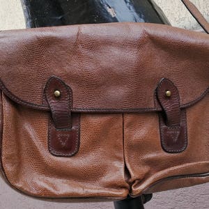 May include: A brown leather satchel bag with a flap closure and two front pockets. The bag has a textured leather finish and a strap.