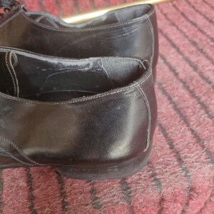 May include: Close-up of a pair of black leather dress shoes. The shoes have a classic design with laces and a low heel. The leather appears worn, with visible creases and wear marks.