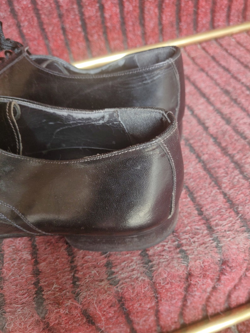 May include: Close-up of a pair of black leather dress shoes. The shoes have a classic design with laces and a low heel. The leather appears worn, with visible creases and wear marks.