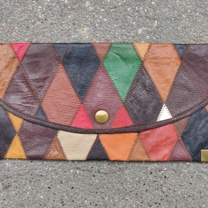 Op de afbeelding: Een bruine leren clutch met een ruitpatroon. De clutch is gemaakt van verschillende gekleurde lederen stukken die aan elkaar zijn genaaid. De clutch heeft een gouden knoopsluiting en gouden hoekaccenten.