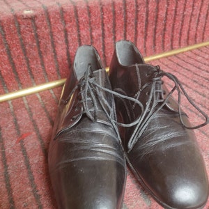 May include: A pair of black leather dress shoes with laces. The shoes are placed on a red carpeted staircase with gold-colored metal accents. The shoes have a classic, formal design.