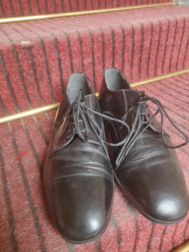 May include: A pair of black leather dress shoes with laces. The shoes are placed on a red carpeted staircase with gold-colored metal accents. The shoes have a classic, formal design.