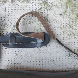 May include: A black leather camera strap with a brown leather adjustment strap. The strap has a metal buckle.