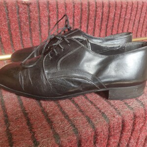 May include: A pair of black leather dress shoes with laces. The shoes have a classic design with a rounded toe and a low heel. The shoes are sitting on a red carpet.